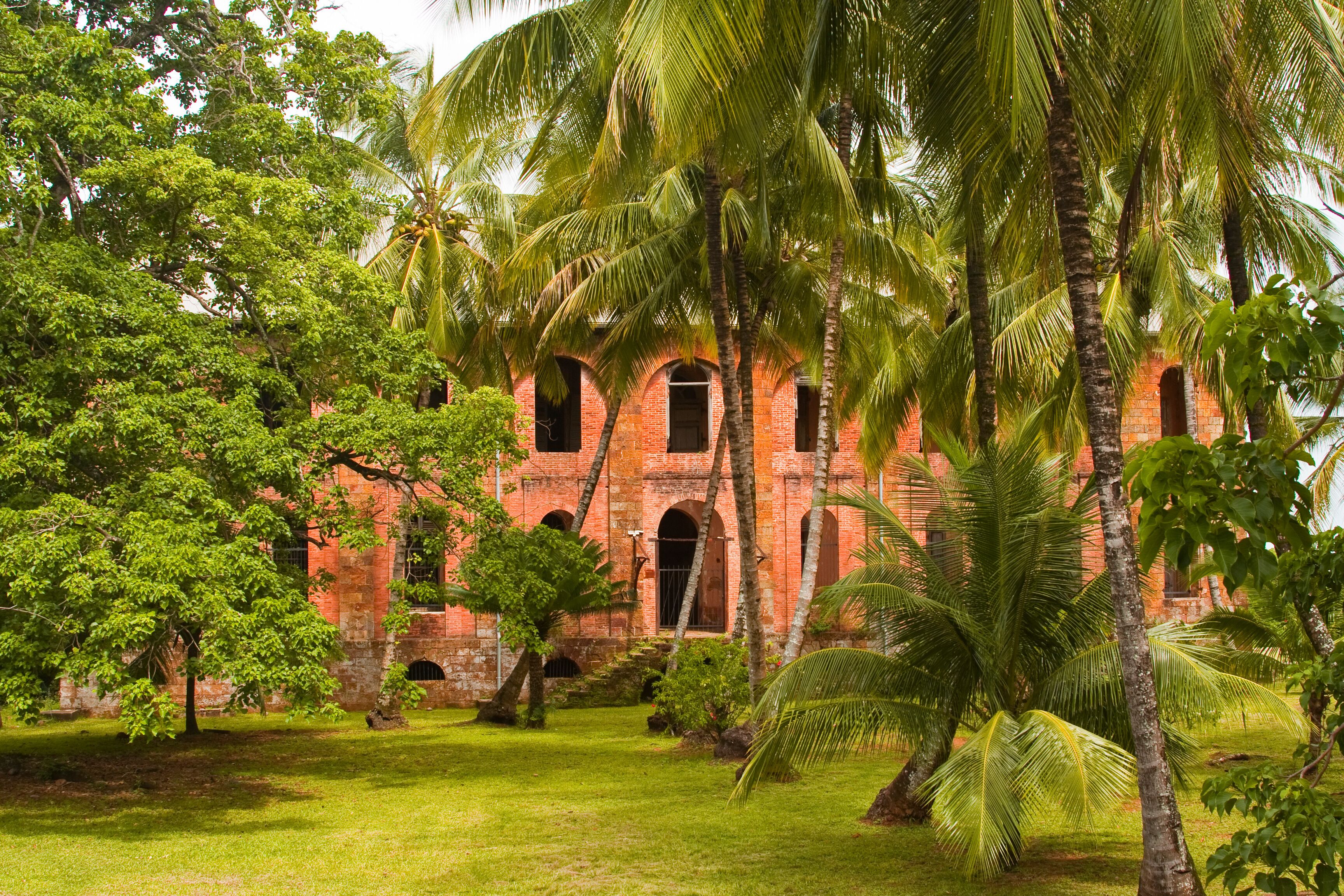 A historic red brick building with arched windows, surrounded by lush greenery and palm trees.
