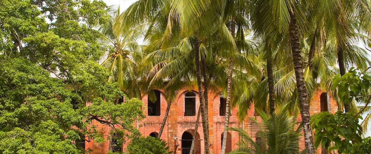 A historic red brick building with arched windows, surrounded by lush greenery and palm trees.