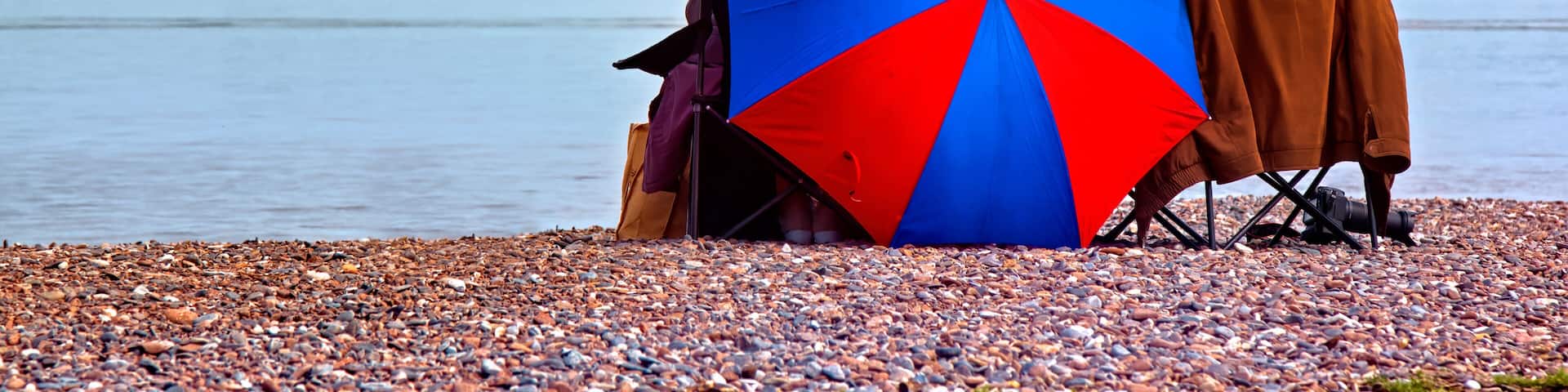 A pebbled beach with a large umbrella and a person in a brown jacket.