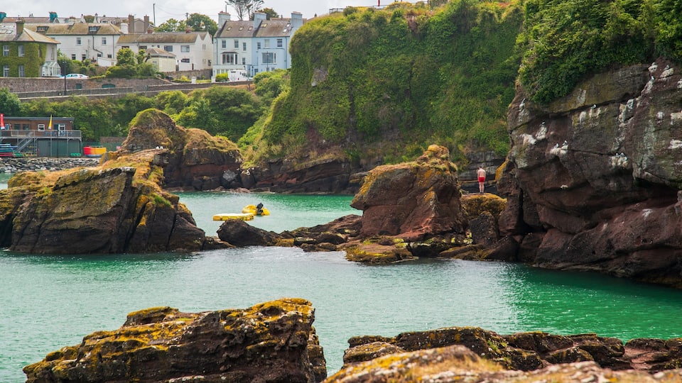 A coastal scene with rocky outcrops, clear turquoise water, and buildings in the background.
