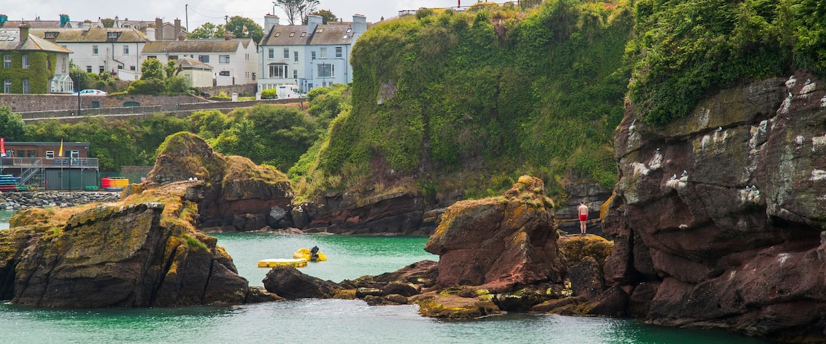 A coastal scene with rocky outcrops, clear turquoise water, and buildings in the background.