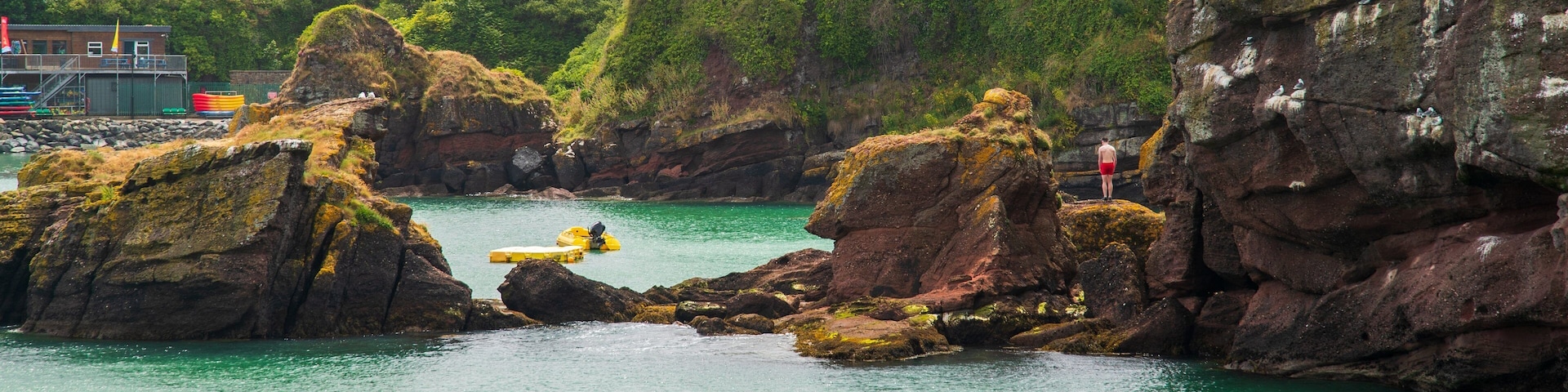 A coastal scene with rocky outcrops, clear turquoise water, and buildings in the background.
