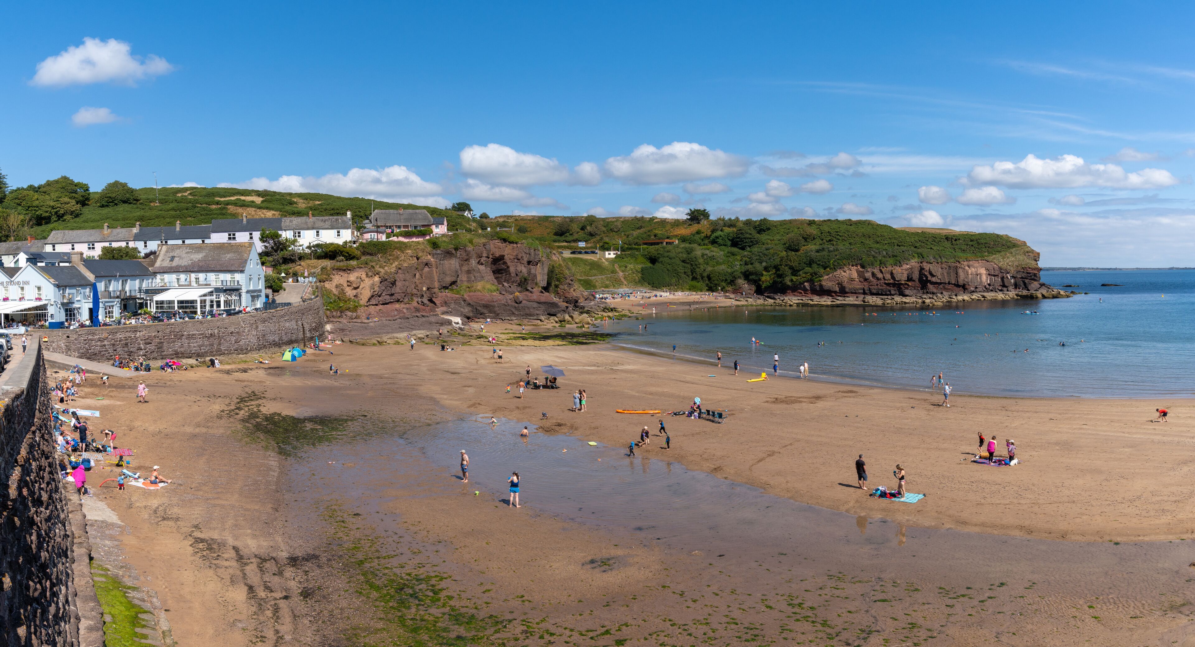 A coastal beach with people sunbathing and swimming, a rocky cliff, and a building with a blue roof.