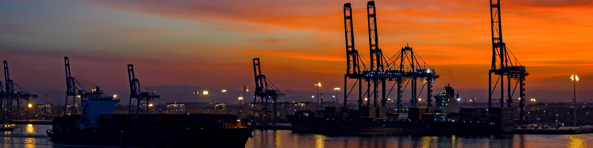 Un puerto al atardecer con buques de carga y grúas silueteadas contra un cielo colorido.