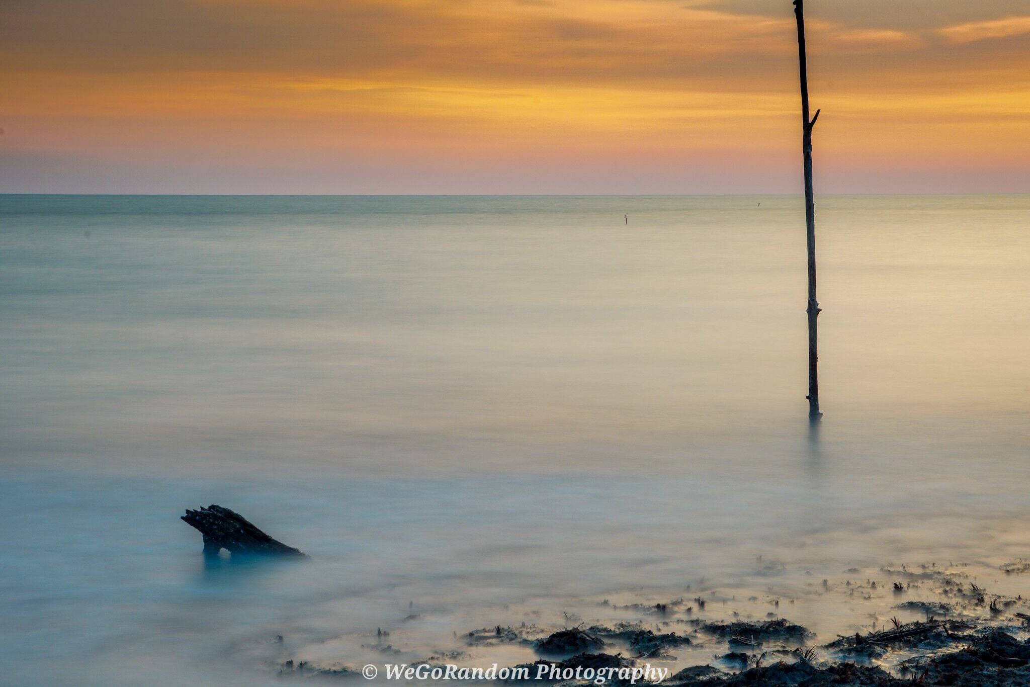 A calm sea at sunset with a single tall pole in the water and a small boat on the shore.