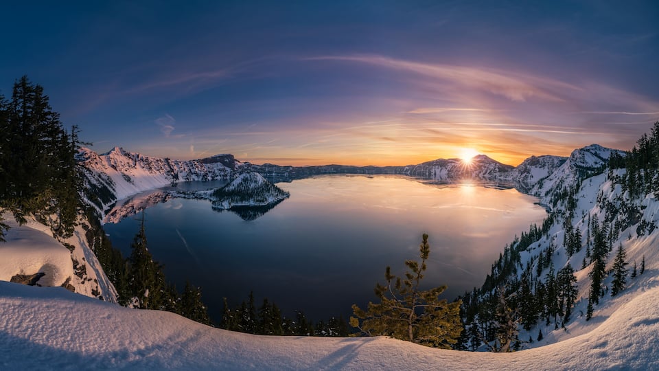A snow-covered mountain landscape with a lake and sunset.