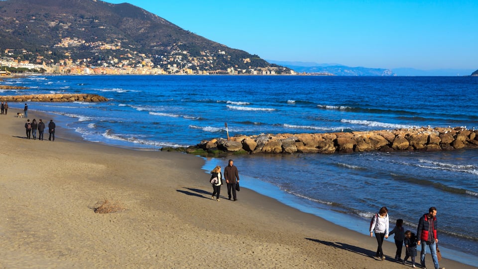 A beach with people walking, rocky breakwaters, and a mountainous backdrop.