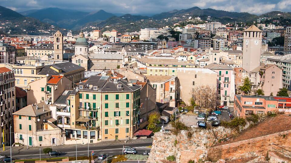 A cityscape with historic buildings, a prominent clock tower, and a mountainous backdrop.