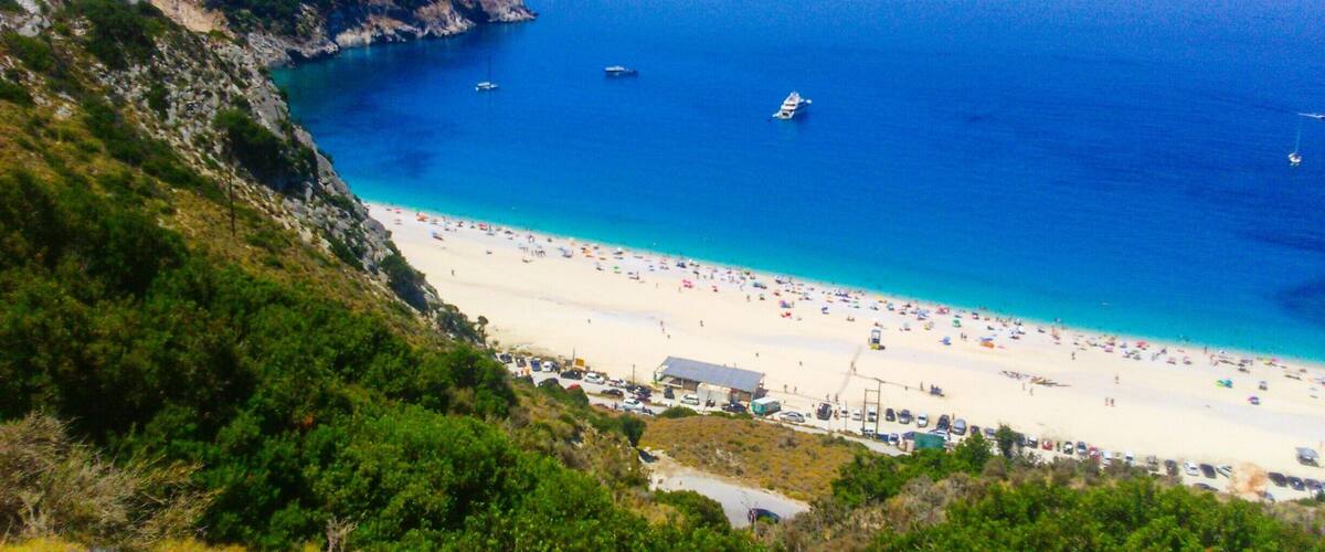 A coastal beach with clear blue waters, boats, and a hilly landscape.