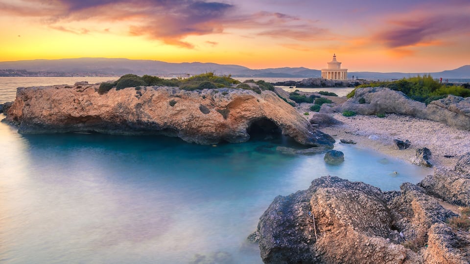 A coastal landscape with a rock formation, clear blue waters, and a distant building.