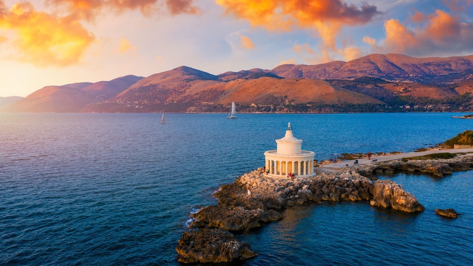 A coastal scene with a white lighthouse on a rocky outcrop, a sailboat in the distance, and mountains in the background.
