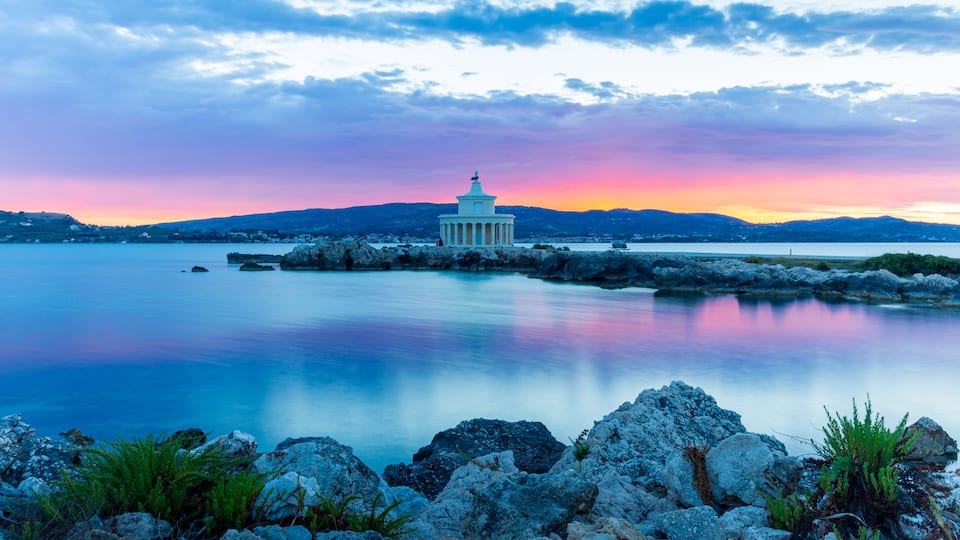 A coastal landscape with a white building on a rocky outcrop, calm water, and a sunset sky.