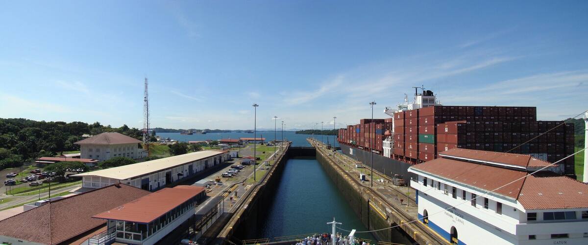 A canal with a lock system, a large cargo ship, and a white building with "PANAMA CANAL" written on it.