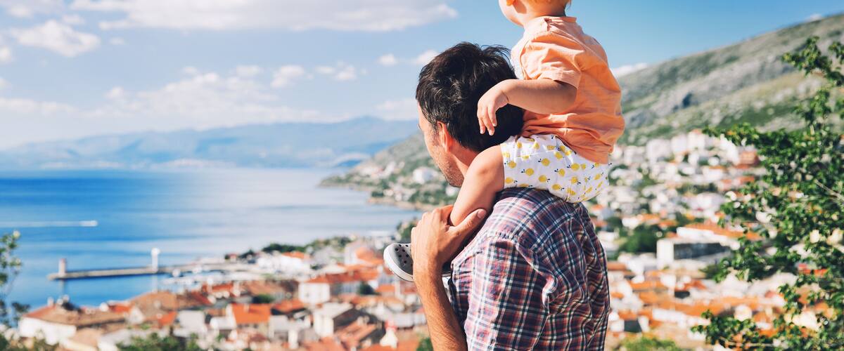 A man carrying a child, overlooking a coastal town with red-roofed buildings and a clear blue sky.