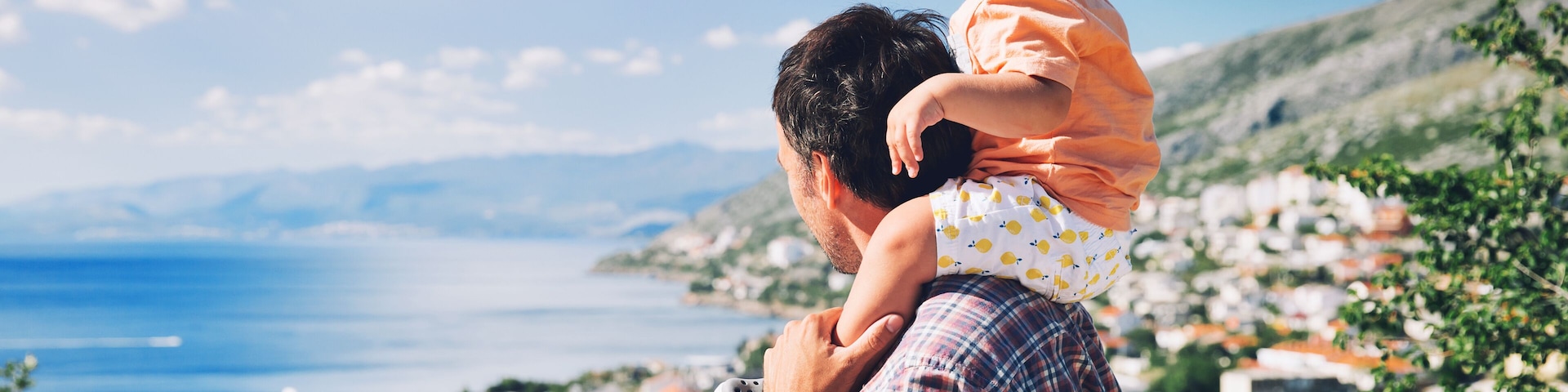 A man carrying a child, overlooking a coastal town with red-roofed buildings and a clear blue sky.