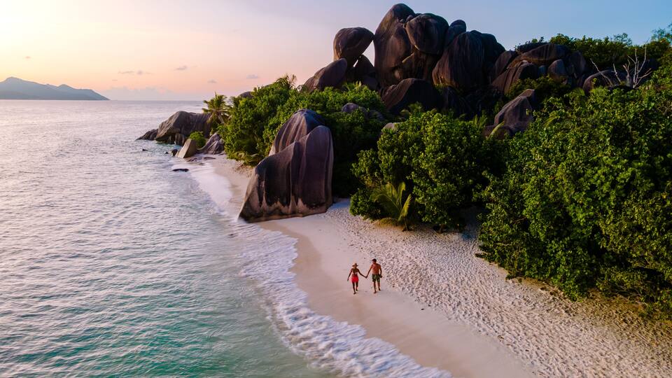 A beach with large boulders, palm trees, and a couple walking on the sand.