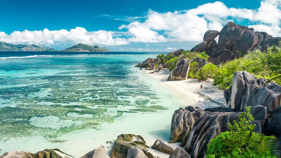 A tropical beach with clear turquoise waters, large boulders, and lush greenery.