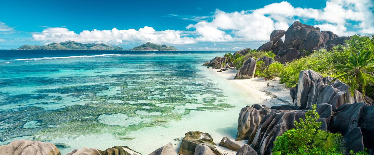 A tropical beach with clear turquoise waters, large boulders, and lush greenery.