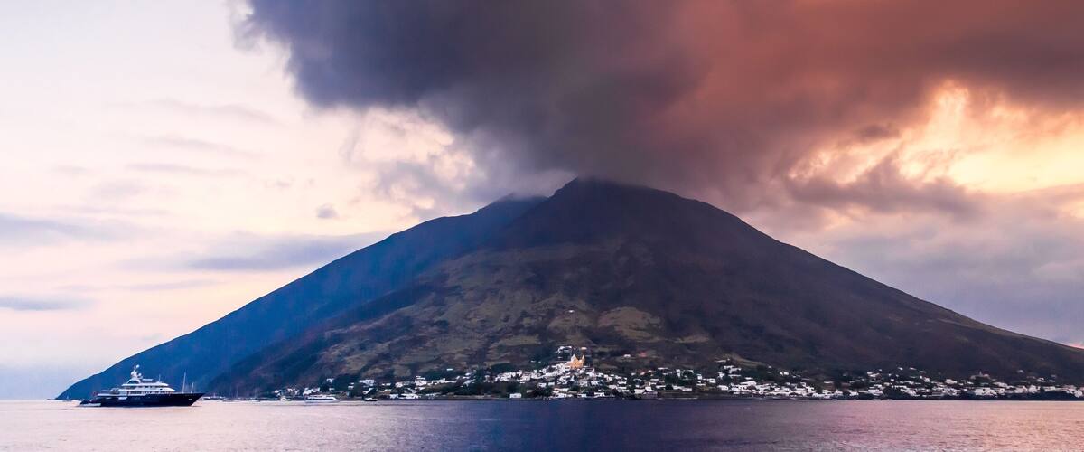 A large volcano with a town at its base and a ship in the foreground.