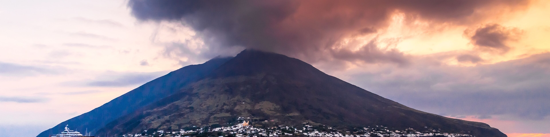 A large volcano with a town at its base and a ship in the foreground.