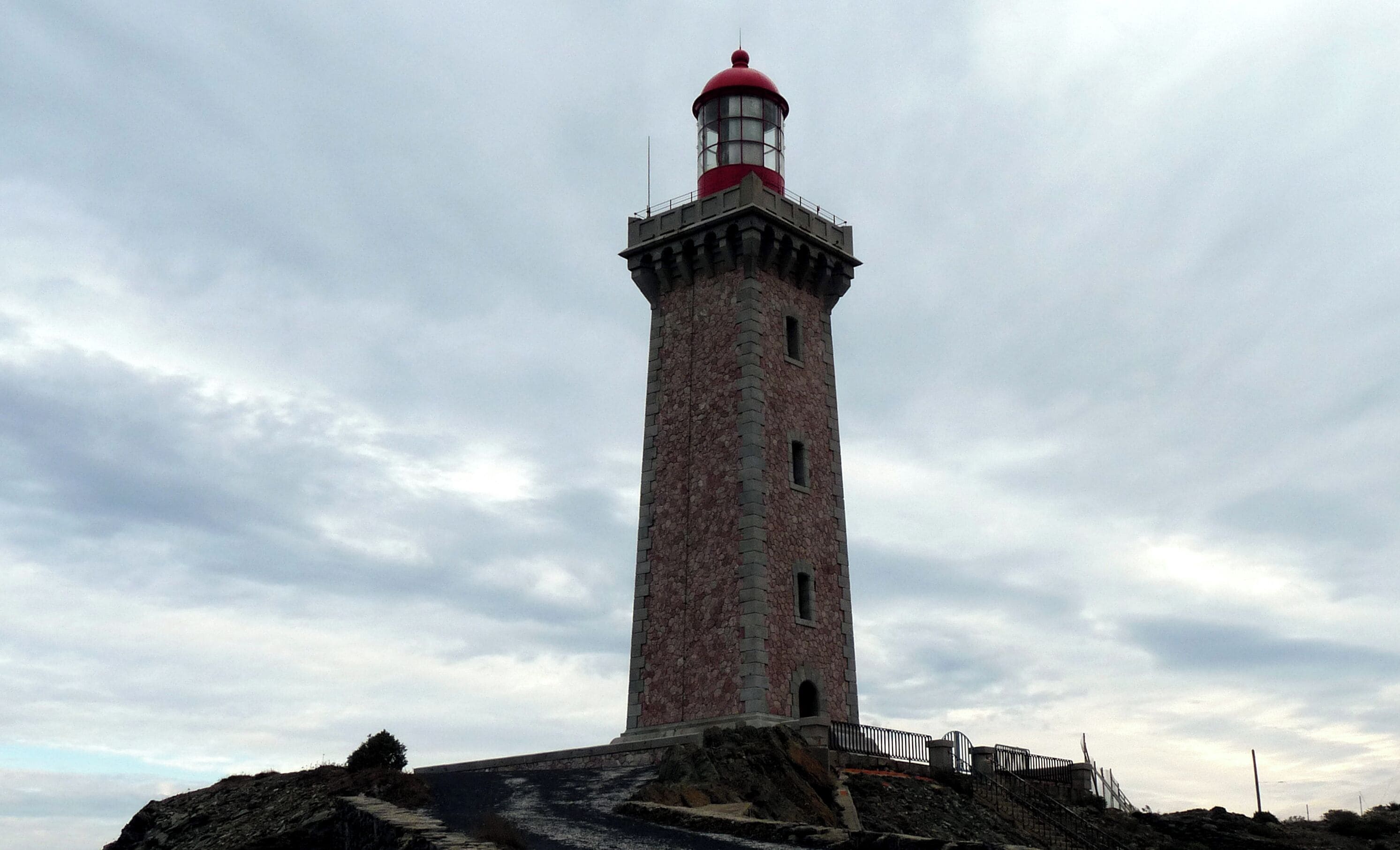 A tall lighthouse with a red top, constructed from pinkish stones, standing on a rocky outcrop with a cloudy sky in the background.