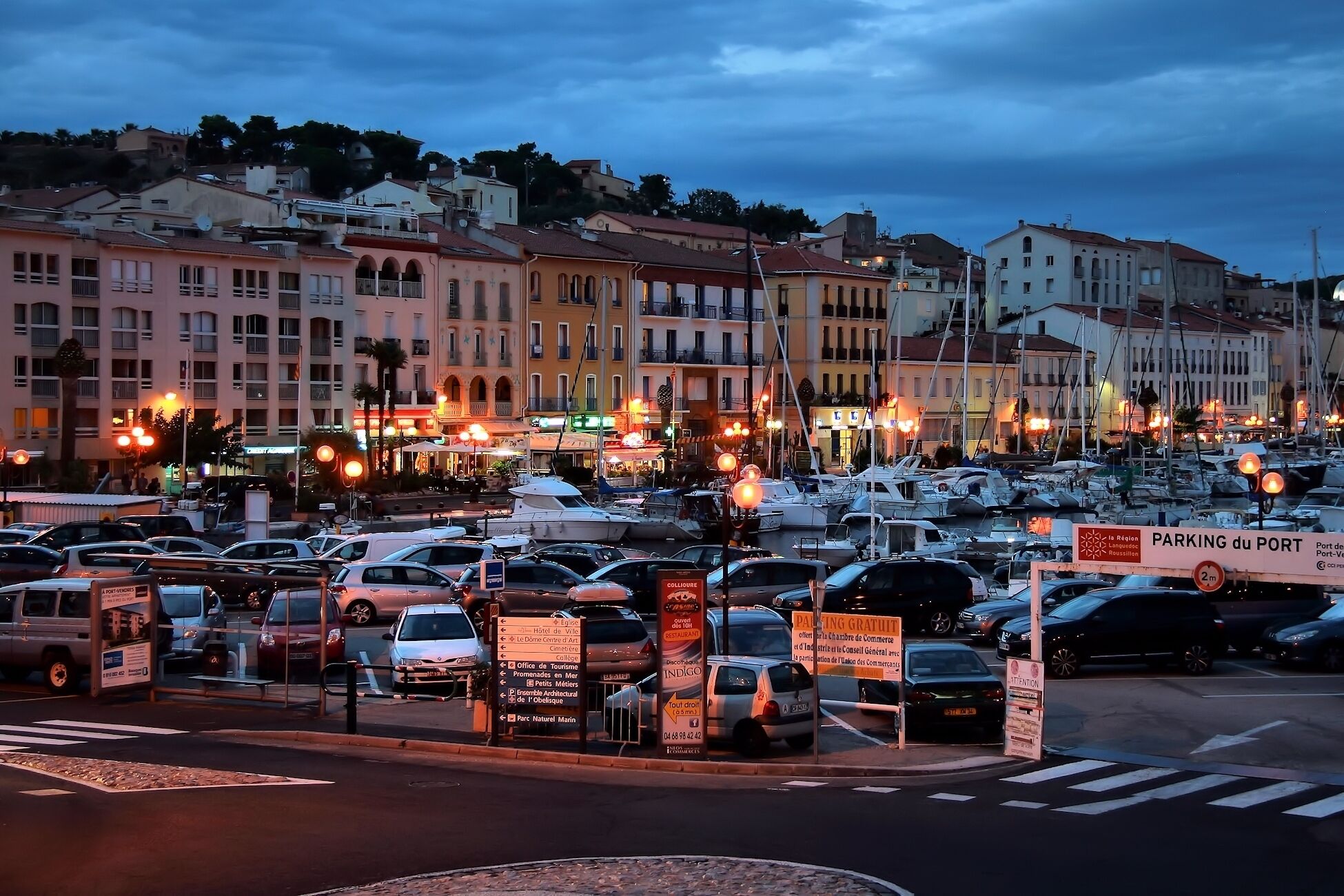 A marina with boats docked, surrounded by buildings and a busy street at dusk.
