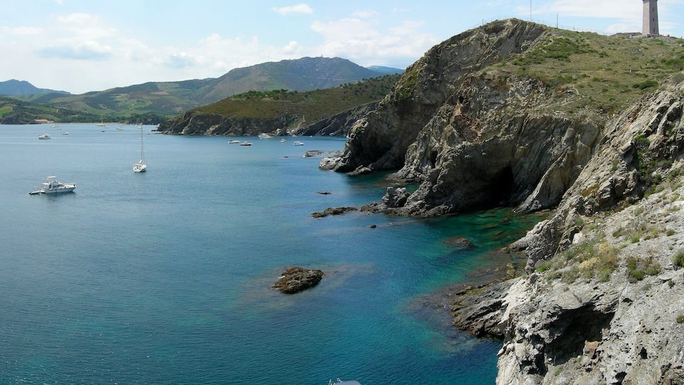 A coastal landscape with a lighthouse, rocky cliffs, and boats on clear blue waters.