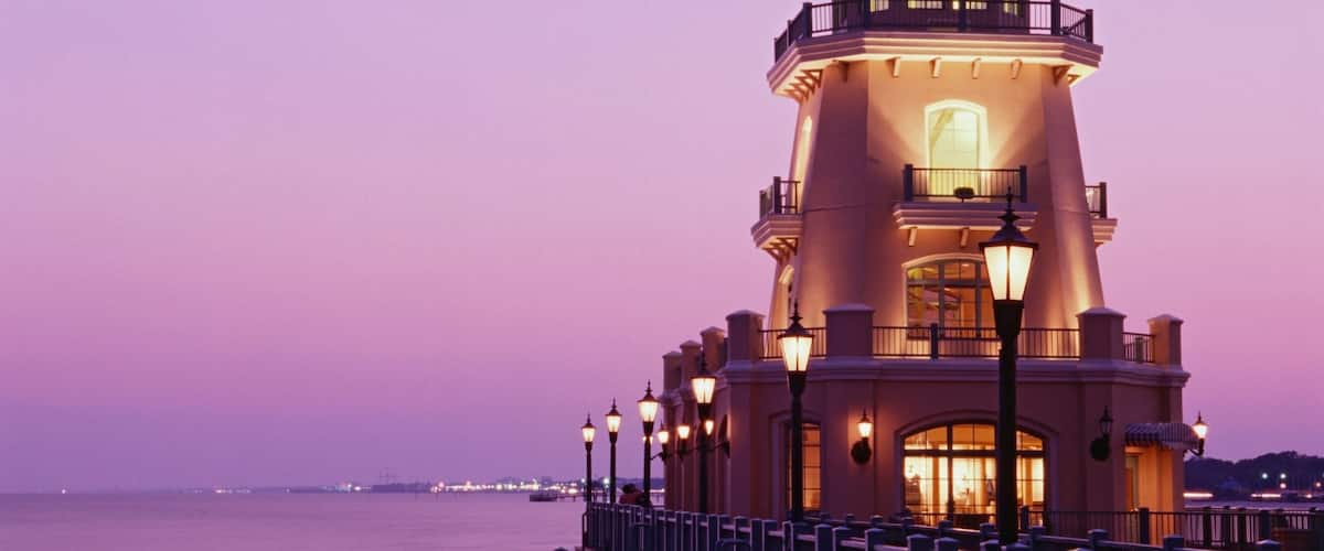 Un faro al anochecer con un cielo morado, un mar tranquilo y un paisaje urbano distante.