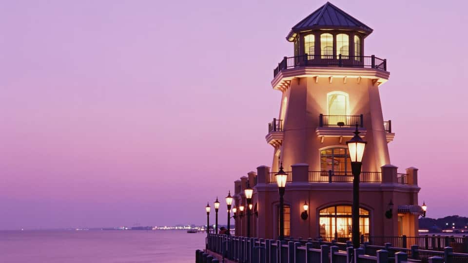 A lighthouse at dusk with a purple sky, a calm sea, and a distant cityscape.