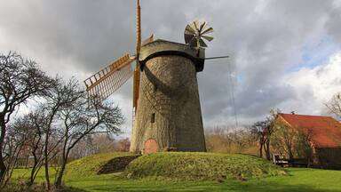 A traditional windmill with a wooden structure and a wind turbine on top, surrounded by a grassy landscape and trees.