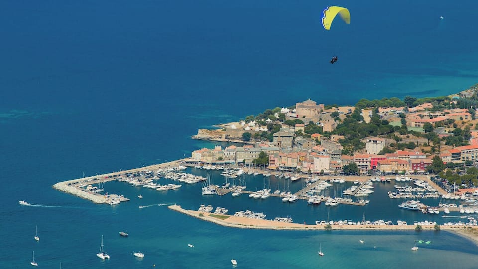 A coastal town with a marina filled with boats, a person paragliding, and historic buildings on a hillside.