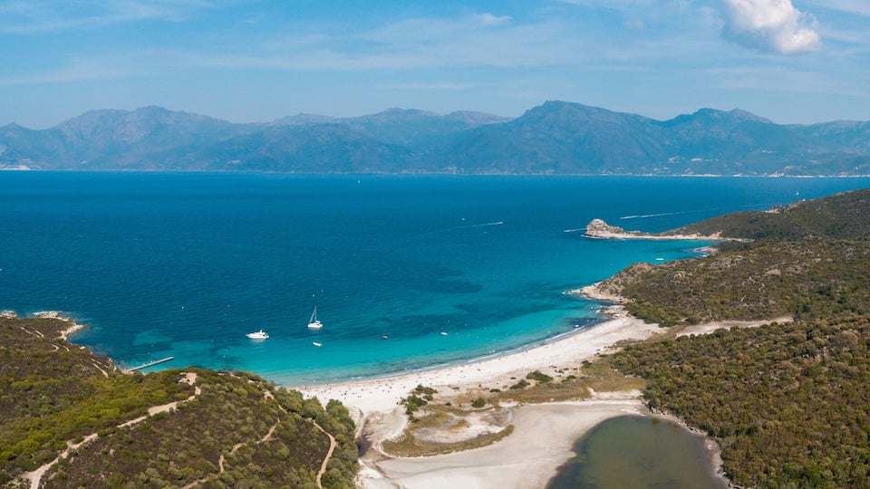 A coastal landscape with a beach, clear blue sea, and distant mountains.