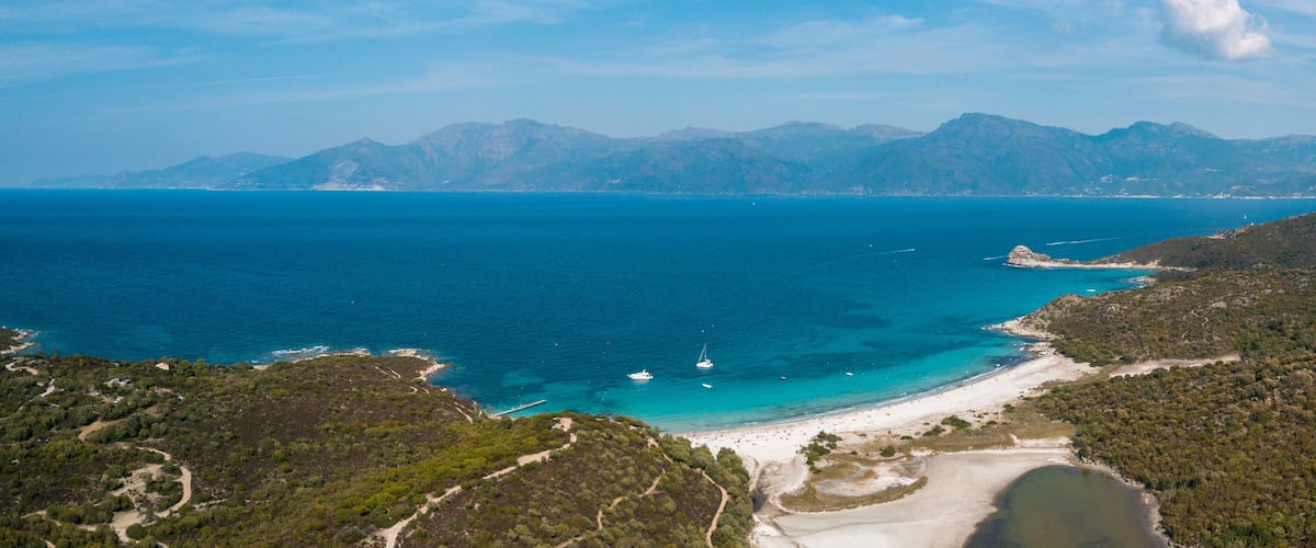 A coastal landscape with a beach, clear blue sea, and distant mountains.
