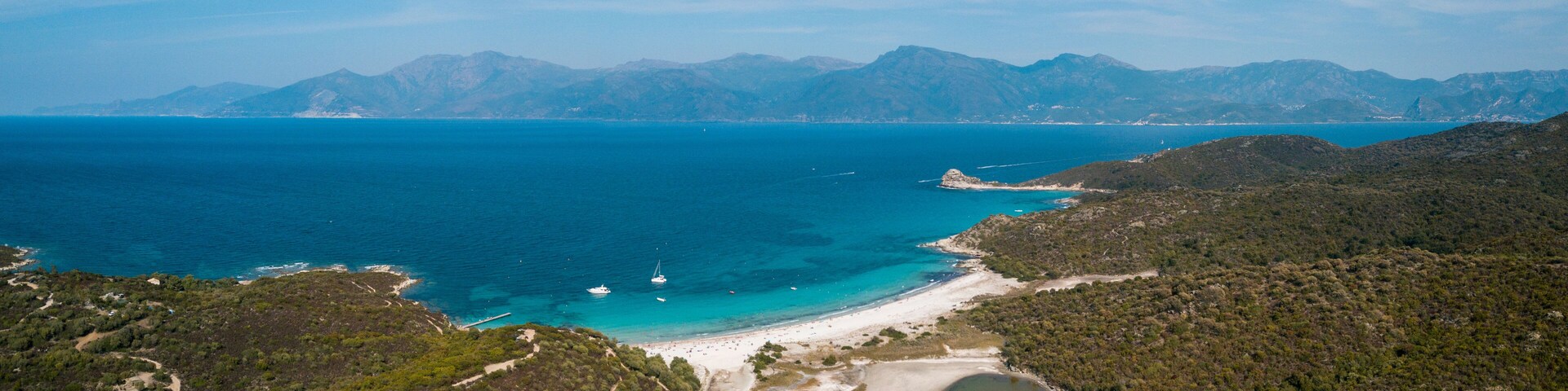A coastal landscape with a beach, clear blue sea, and distant mountains.