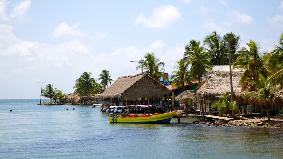 A tropical beach with thatched-roof huts, palm trees, and a yellow boat docked near the shore.