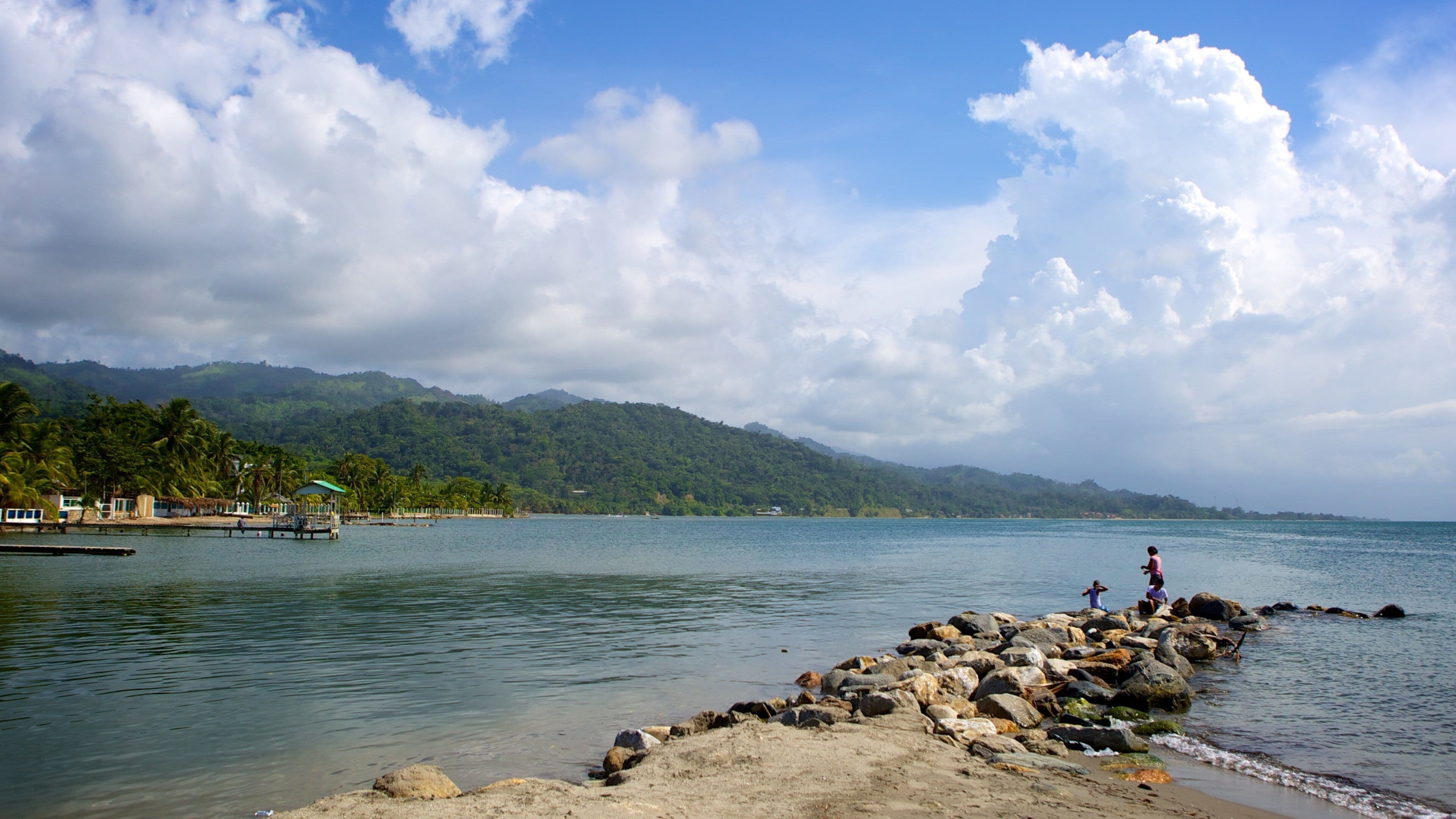 A coastal scene with a rocky breakwater, calm water, and a backdrop of green hills under a partly cloudy sky.