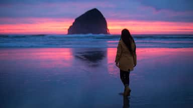 Uma pessoa caminhando na praia com uma grande formação rochosa ao longe durante o pôr do sol.
