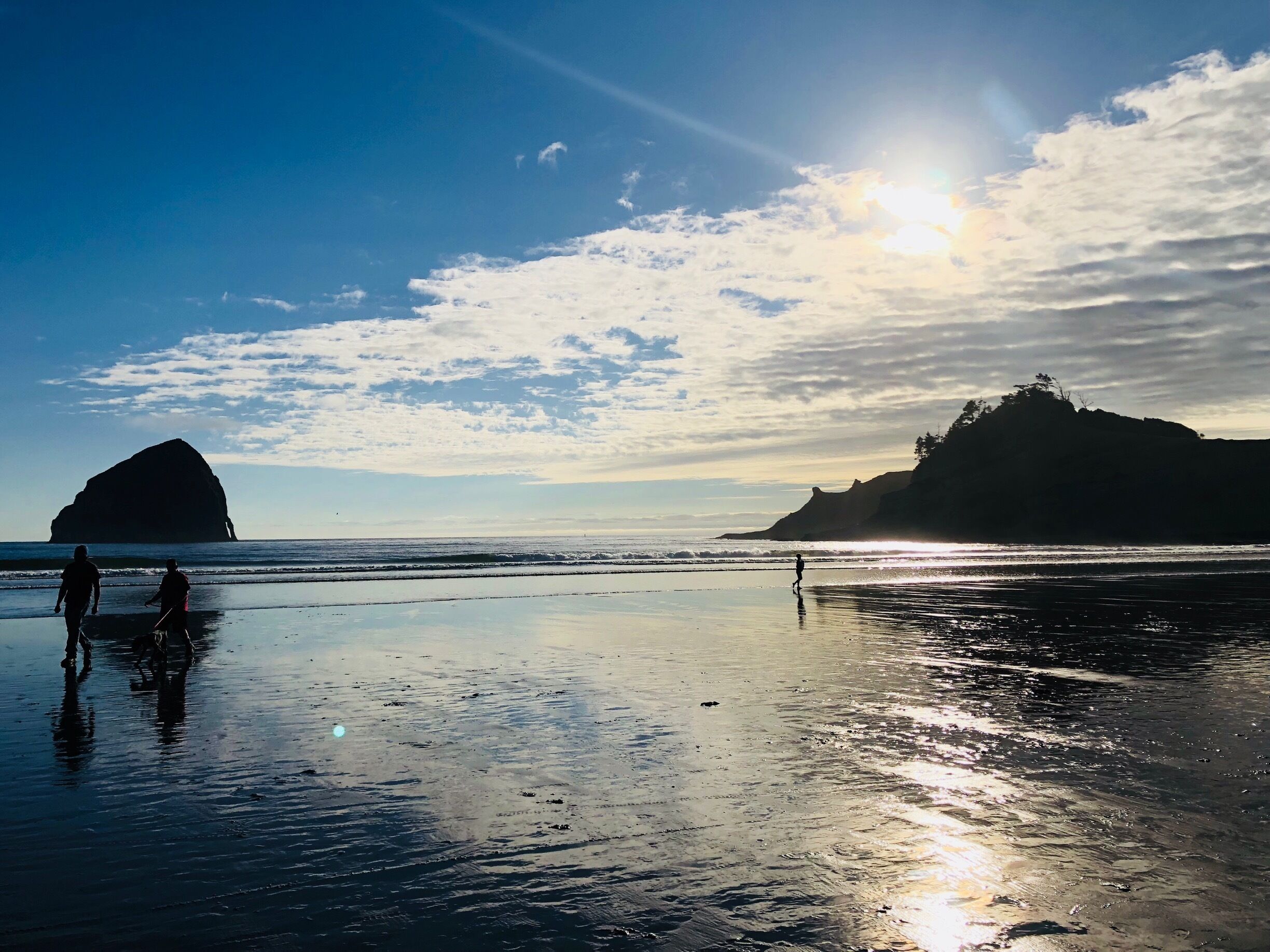 A beach with a large rock formation in the distance, a person walking, and the sun setting over the water.