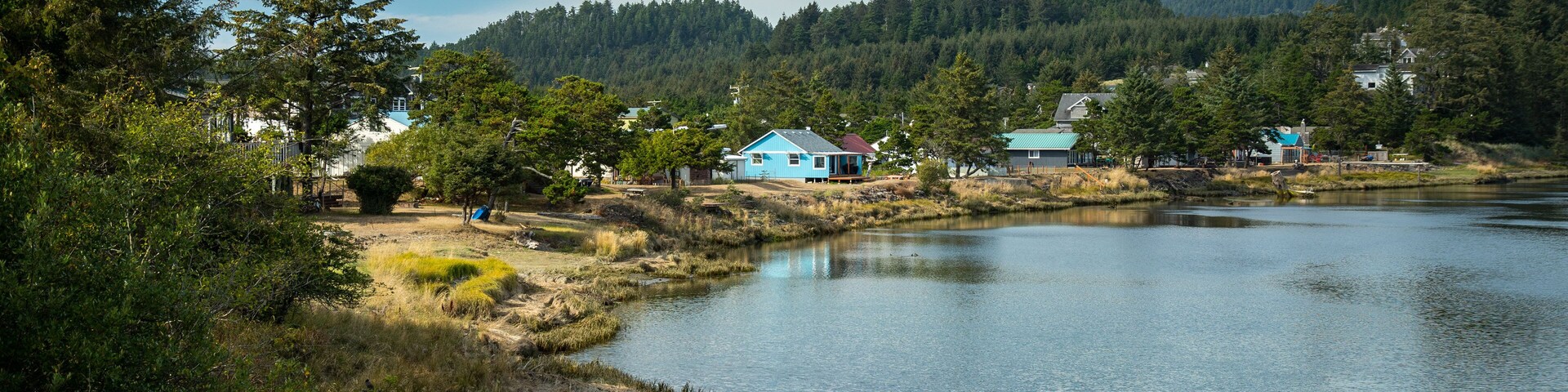A calm lake with houses on the shore, surrounded by dense forest and hills.