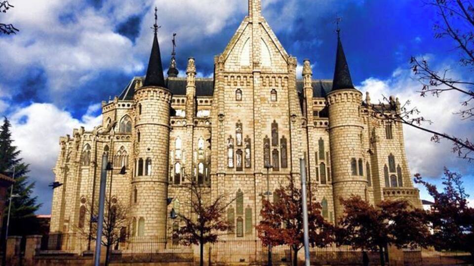 A large, historic stone building with multiple towers and spires, surrounded by a paved area with benches.