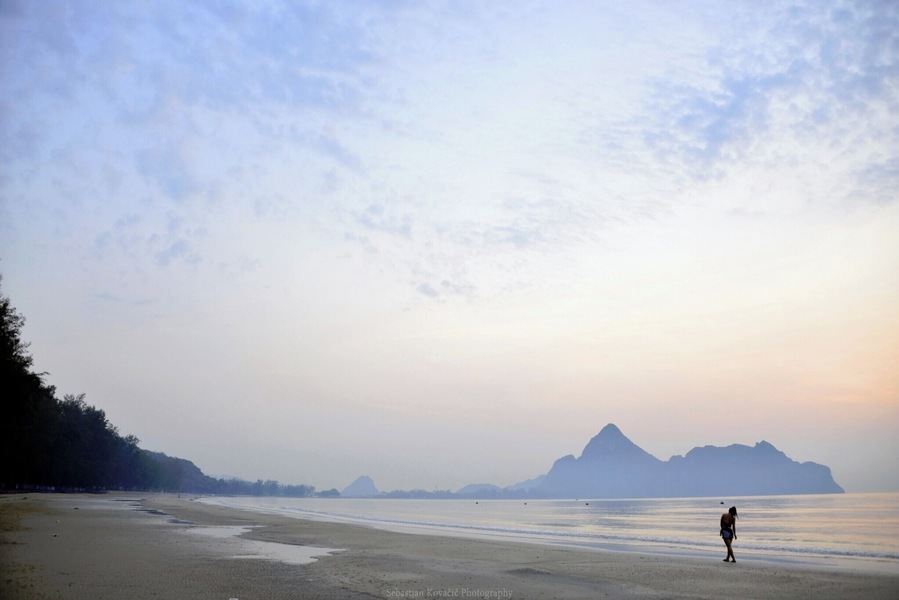 Uma praia com uma pessoa caminhando, águas calmas e montanhas ao longe.