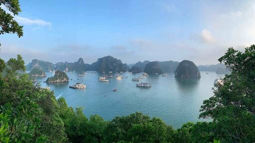 A coastal scene with numerous boats on calm waters, surrounded by lush green islands and rocky formations.