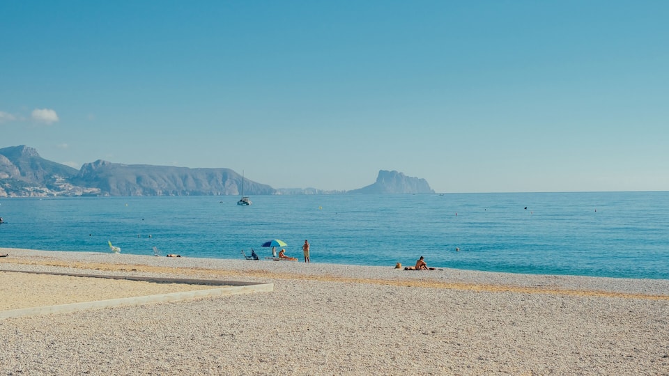 A pebble beach with people sunbathing and swimming, a clear blue sea, and distant mountains.