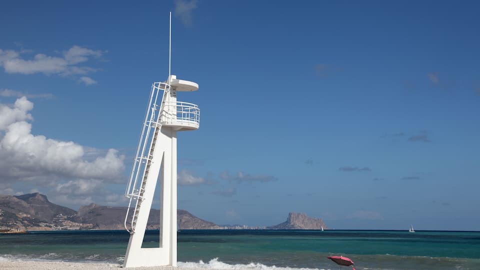 A white lighthouse on a pebbly beach with clear blue water and distant mountains.