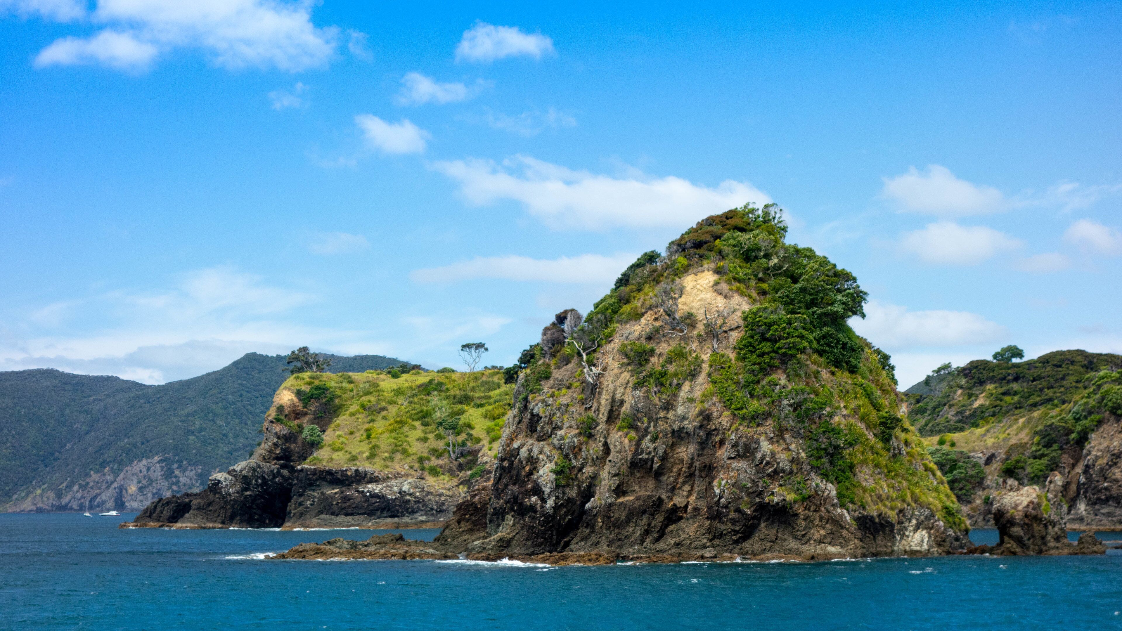A rugged coastal landscape with a prominent rocky island surrounded by clear blue waters and distant mountains.