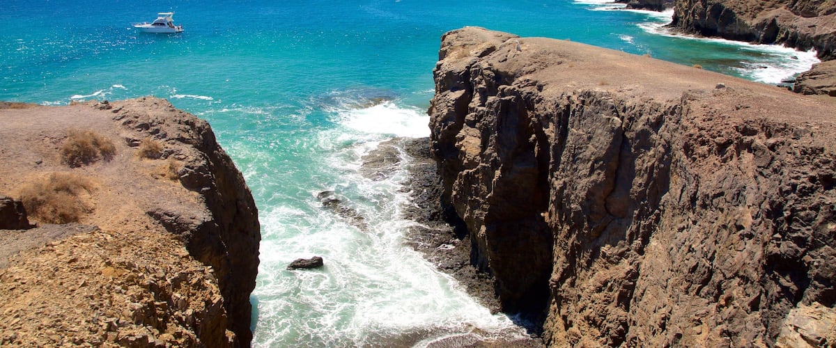 A rocky coastline with clear blue waters, a boat in the distance, and a mountainous horizon.
