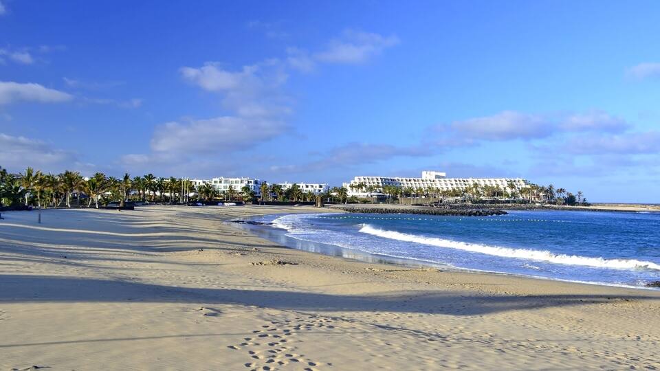 A beach with footprints, palm trees, and a resort in the background.