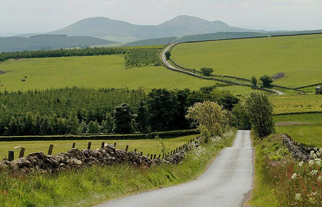 A rural road winding through green fields with stone walls and distant mountains.