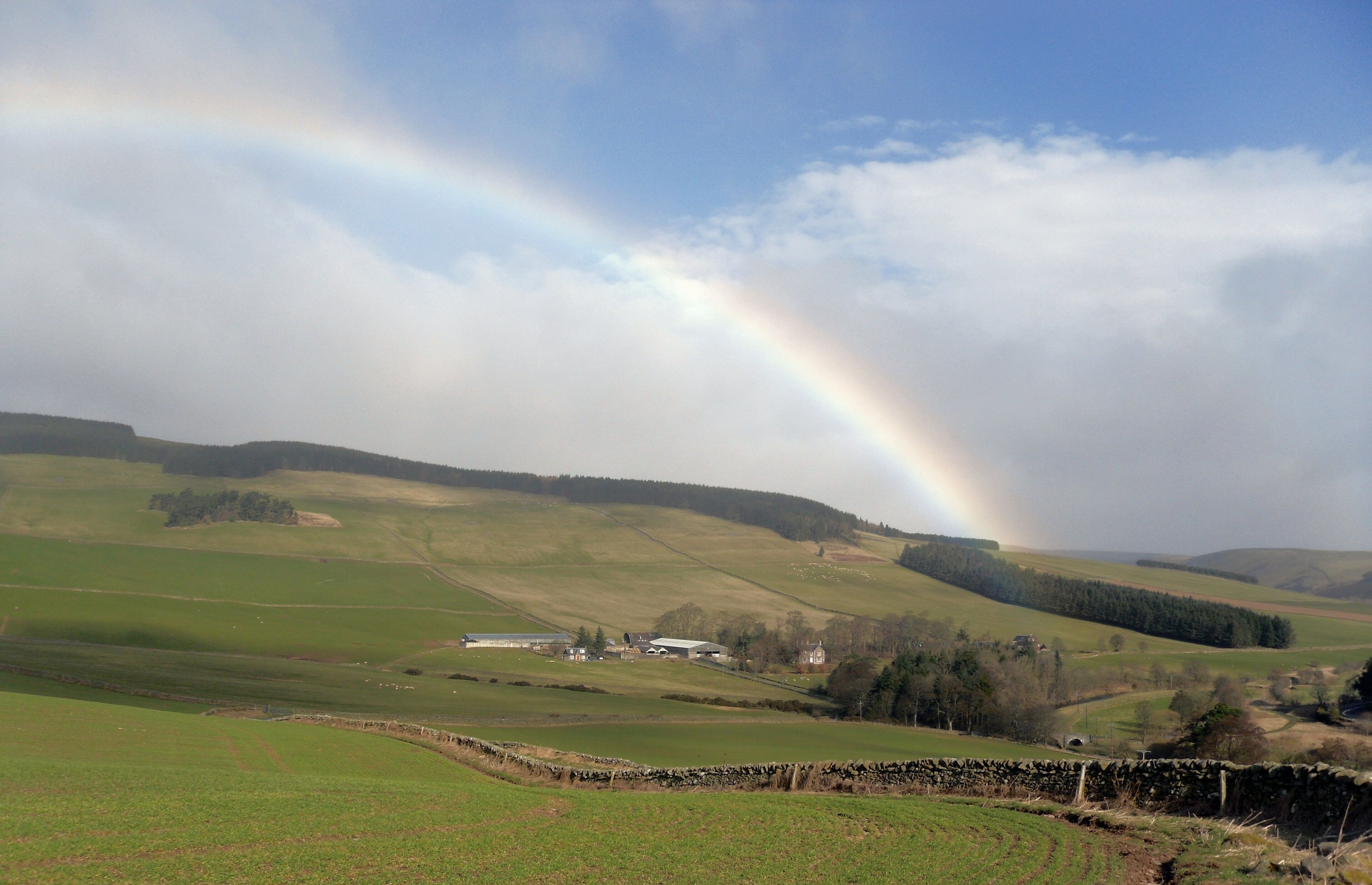 A rural landscape with rolling hills, a rainbow, and scattered buildings.