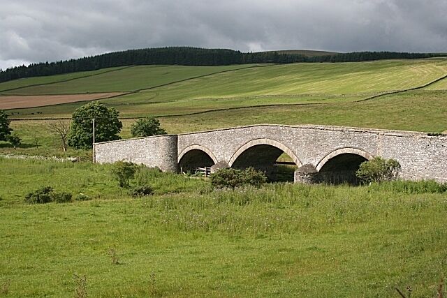 A stone bridge with three arches over a grassy field, surrounded by rolling hills and a cloudy sky.